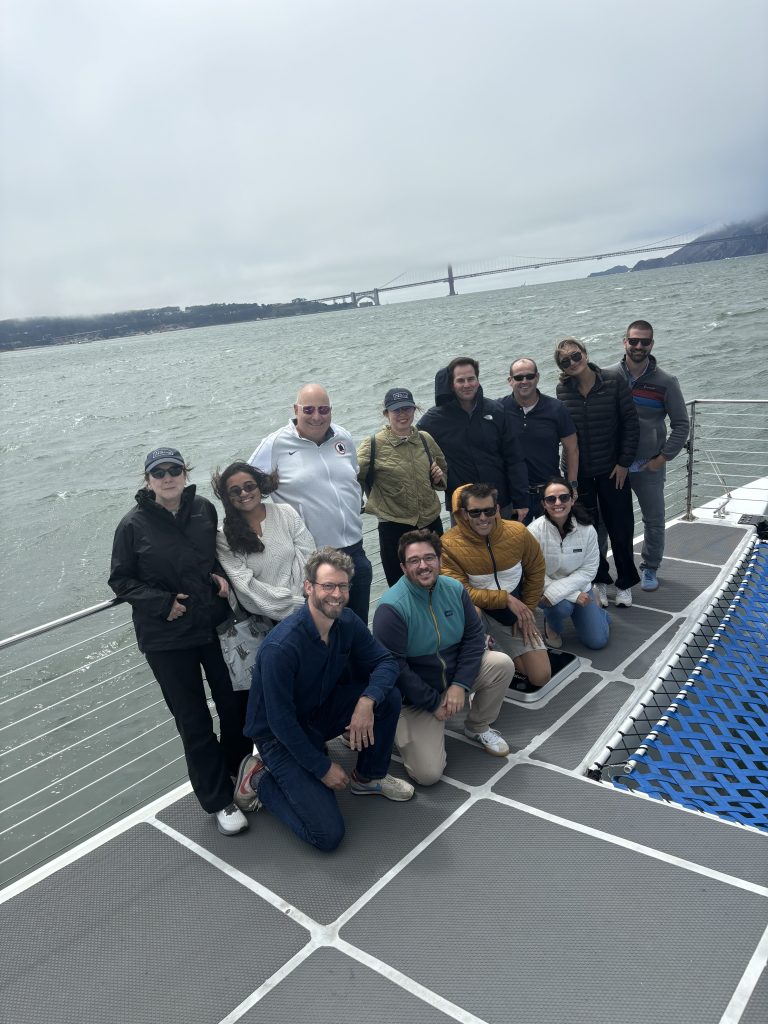 A group of people posing on a boat deck in open water, with the Golden Gate Bridge visible in the background under cloudy skies.
