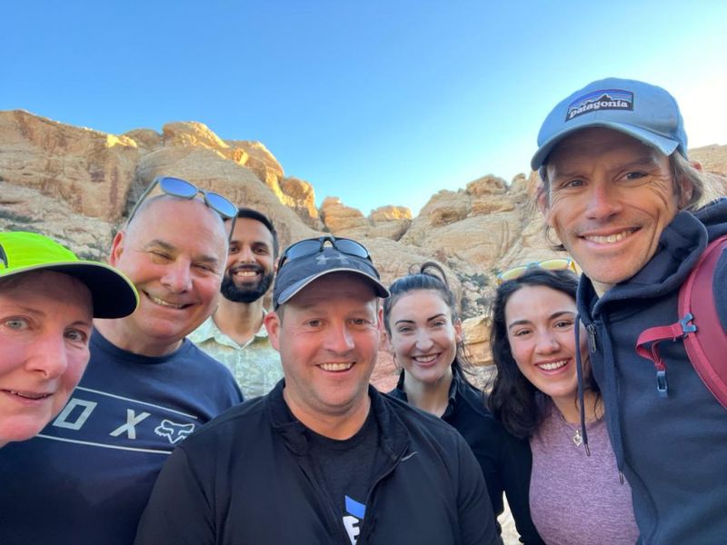 A close-up group selfie featuring seven people smiling at the camera outdoors under a bright blue sky